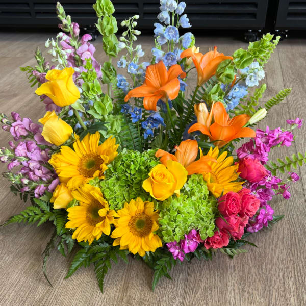 Low arrangement of yellow sunflowers, orange lilies, roses, and mixed bright blooms on a wood floor