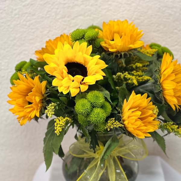 Bouquet of yellow sunflowers and green pom flowers in a glass vase with a yellow ribbon.