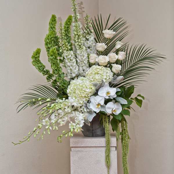 Tall white floral arrangement with orchids, roses, and palm fronds in a gray vase