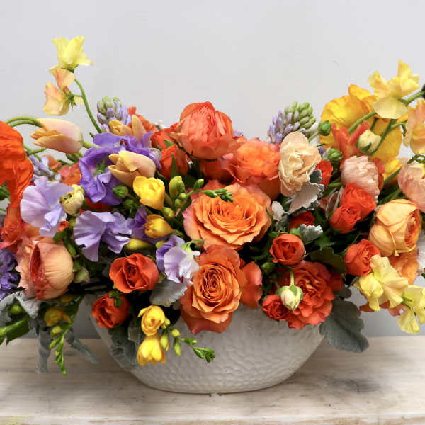 Low ceramic bowl filled with orange roses, ranunculus, and mixed yellow and lavender blooms in a lush arrangement