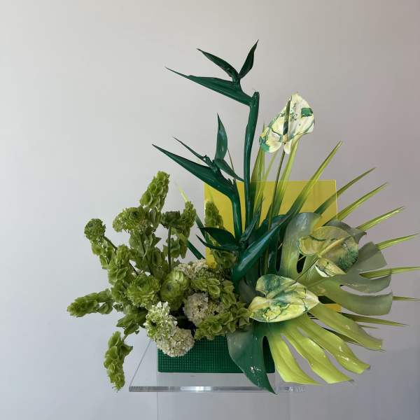 Green floral arrangement with tropical leaves and pale blooms in a square vase