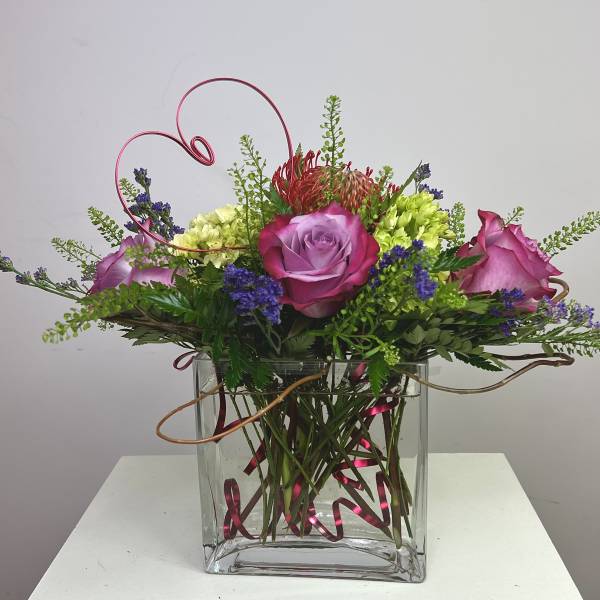Pink and purple roses arranged in a clear square vase with curly wire accents