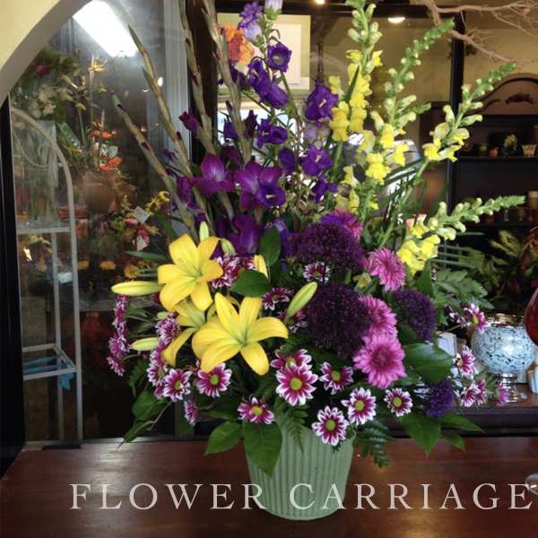 Tall mixed flower arrangement in a green vase with yellow lilies and purple blooms
