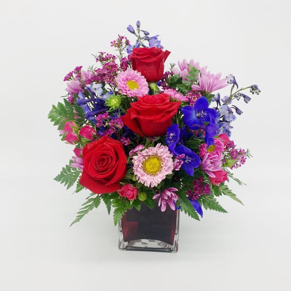 Vibrant mixed bouquet with red roses, pink daisies, and blue flowers in a red glass cube vase