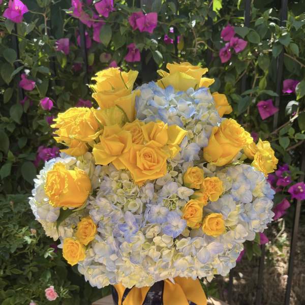 Yellow roses and blue hydrangeas in a dark vase with a yellow ribbon