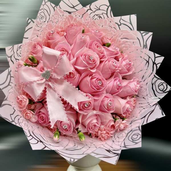 Hand-tied bouquet of pink roses with jeweled centers in pink wrap and bow on a white pedestal vase
