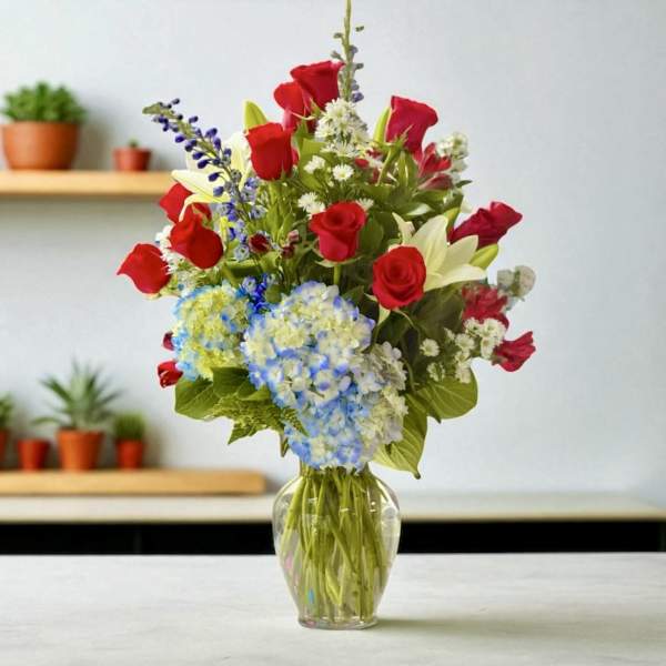 Tall mixed bouquet of red roses, white lilies, blue hydrangeas and small white blooms in a clear glass vase.