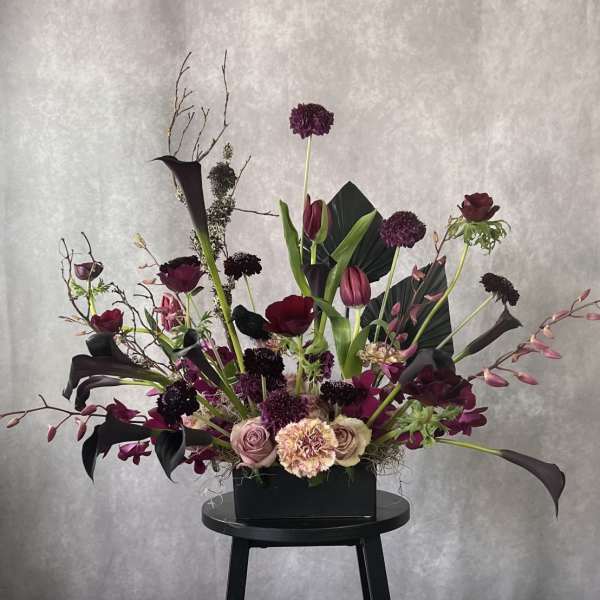 Tall floral arrangement with dark purple blooms in a black rectangular container