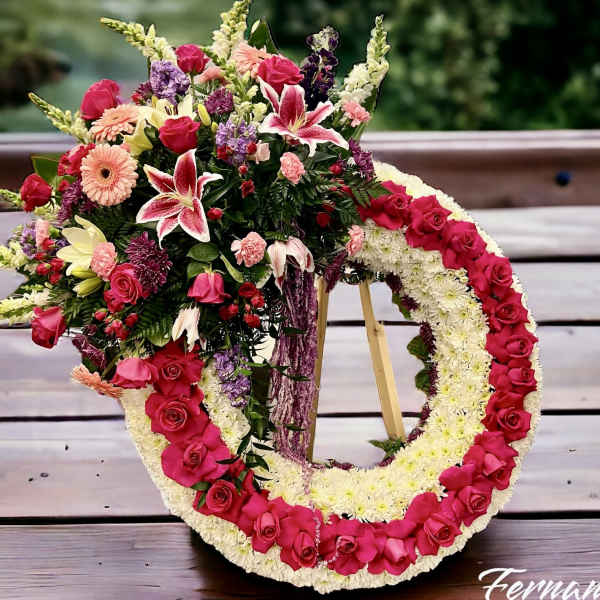 Standing circular wreath of white mums with hot pink roses and mixed pink flowers on a wooden easel.