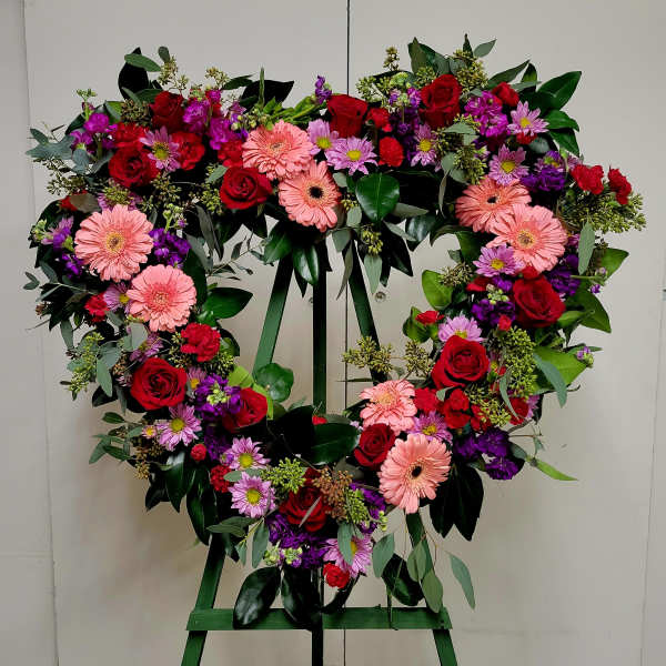 Heart-shaped floral wreath with roses, gerbera daisies, and purple blooms on an easel