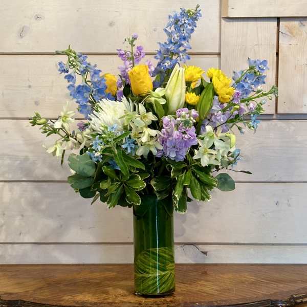 Mixed bouquet of yellow, blue, and lavender flowers in a tall green vase