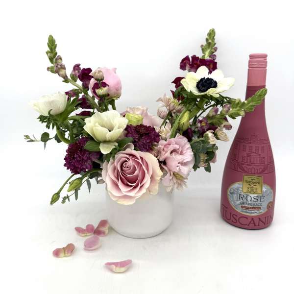 Mixed pink and white flower arrangement in white vase beside pink rosé bottle and scattered petals