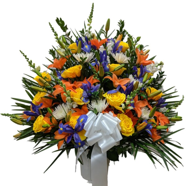 Tall fan-shaped arrangement of yellow roses, orange lilies, blue irises and white flowers with a white bow