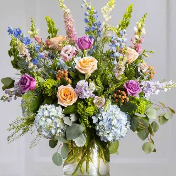 Mixed arrangement of lavender and peach roses, blue hydrangeas, and tall delphinium in a clear glass vase