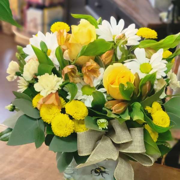 Yellow and white mixed flower arrangement with roses and daisies in a bee-themed glass cube vase with a burlap bow