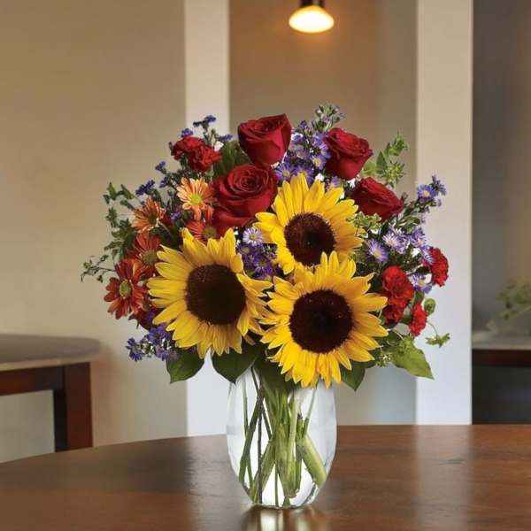 Bouquet of sunflowers, red roses, and purple flowers in a clear glass vase