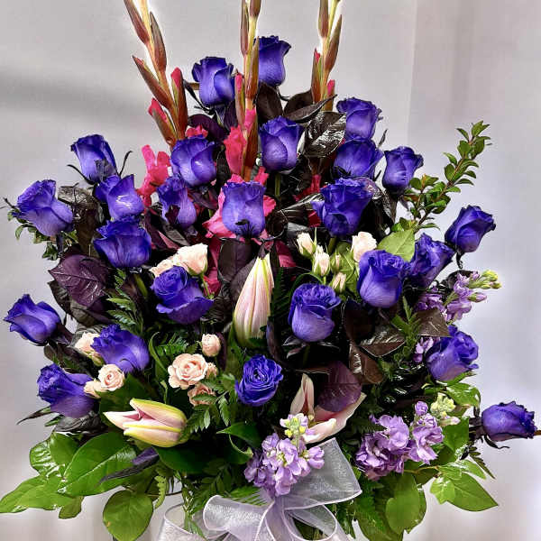 Tall arrangement of purple roses, pink lilies, and mixed blooms in a vase with a sheer ribbon bow