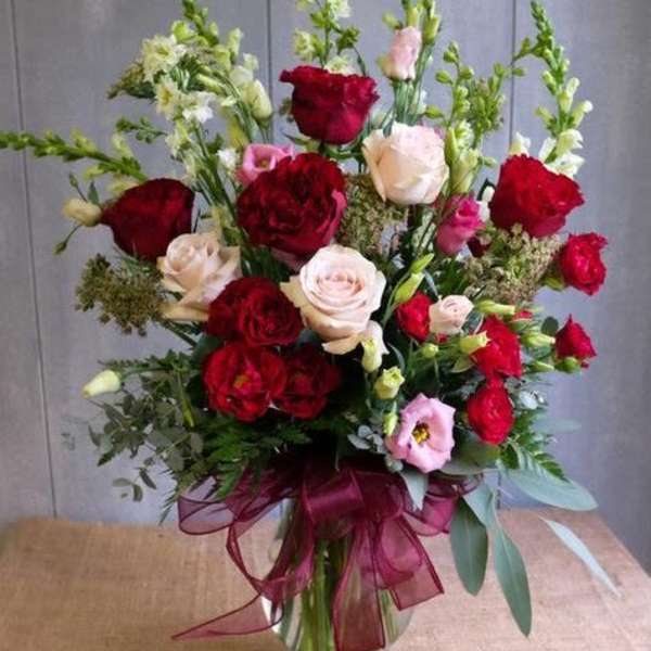 Tall arrangement of red and blush roses with pink blooms and white snapdragons in a glass vase with burgundy bow