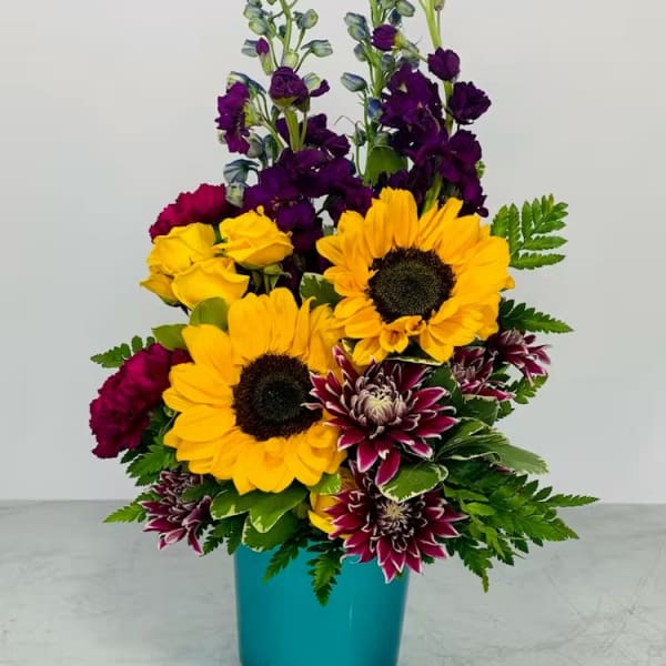 Tall arrangement with sunflowers, purple blooms, and bicolor mums in a turquoise vase