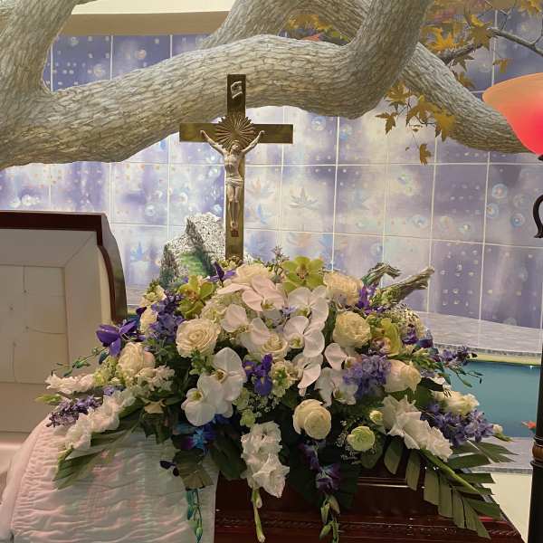 Large funeral flower arrangement with white and purple blooms beneath a crucifix