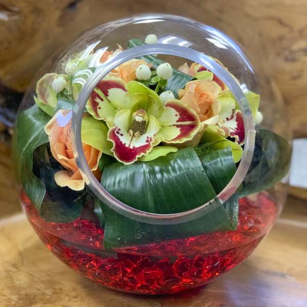 Orchid and peach rose arrangement in a clear glass orb vase with red stones