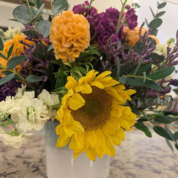 Bright arrangement with a large yellow sunflower, orange and purple blooms in a white vase