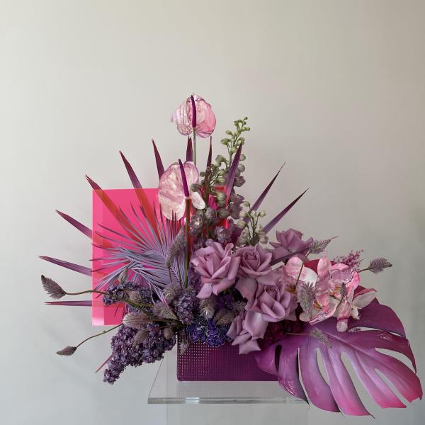 Purple floral arrangement with roses, orchids, and tropical leaves in a pink container