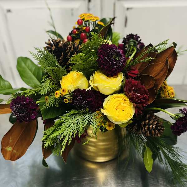 Yellow and burgundy flowers arranged in a gold vase with pinecones and greenery.