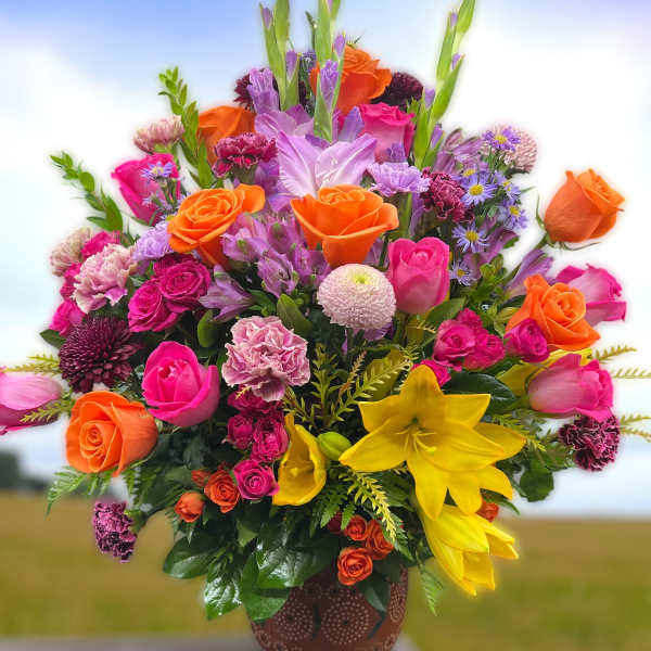 Tall mixed arrangement of pink and orange roses, yellow lilies, and purple accents in a brown pot