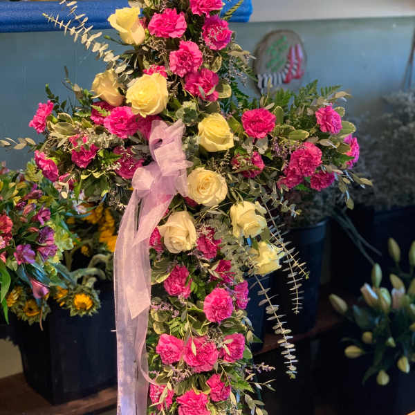 Standing cross spray of hot pink carnations and pale yellow roses with a sheer ribbon bow.
