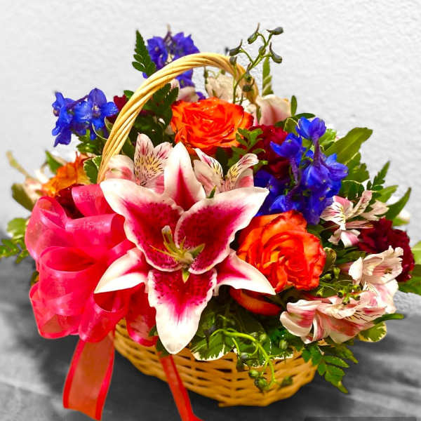Basket arrangement with pink lilies, orange roses, blue flowers, and a pink ribbon
