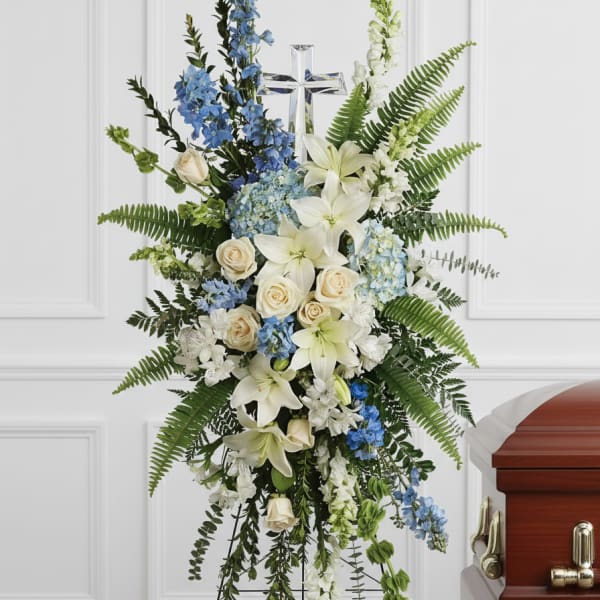 Tall standing spray of white lilies, cream roses, and blue flowers with a cross on an easel