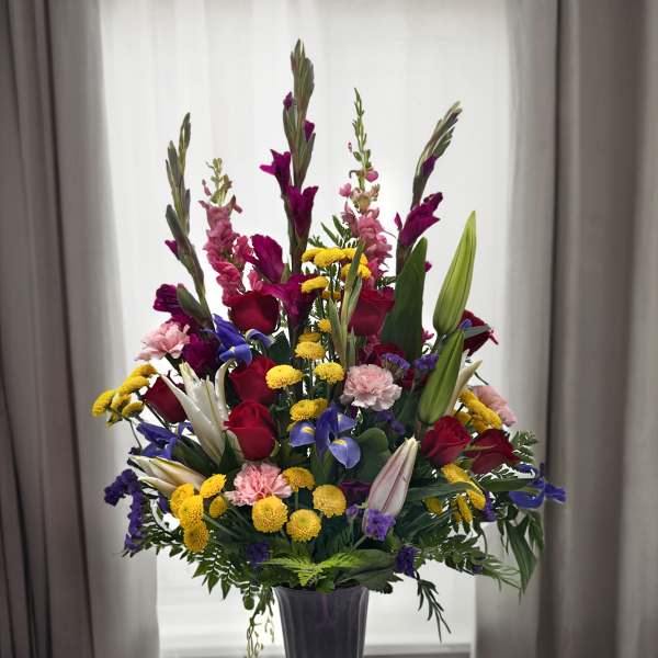 Tall mixed bouquet of roses, lilies, and colorful filler flowers in a purple vase