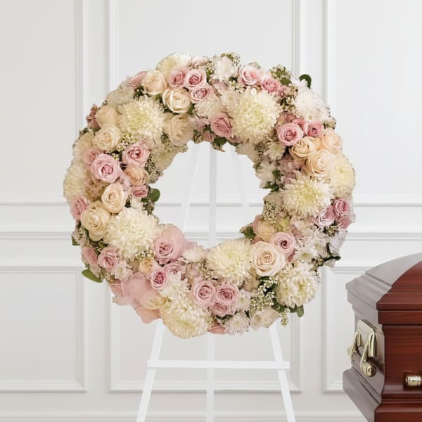 Pastel floral wreath on a white stand beside a wooden casket