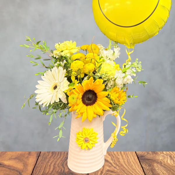 Yellow and white flower arrangement in a pitcher with a yellow smiley balloon