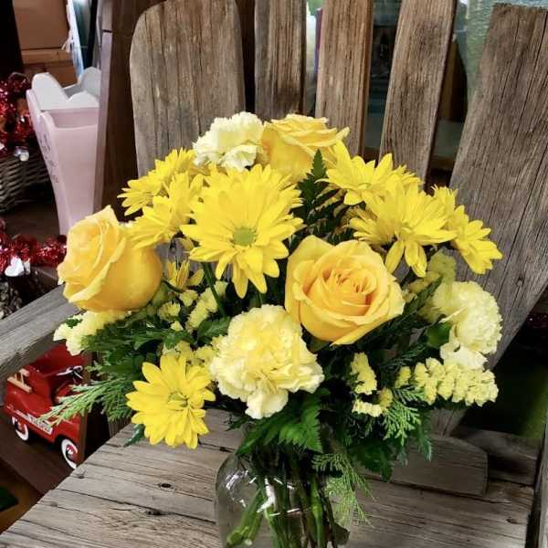 Yellow roses, daisies, and carnations arranged in a clear glass vase on a rustic wooden surface
