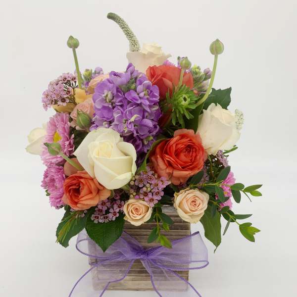 Mixed bouquet of pink, orange, and white roses with purple blooms in a wood box with lavender bow.