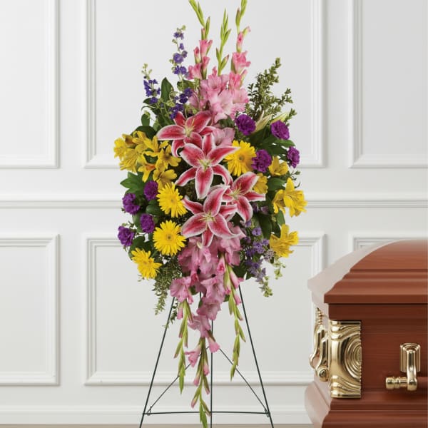 Tall standing spray of pink lilies, yellow daisies, and purple flowers on an easel beside a wooden casket