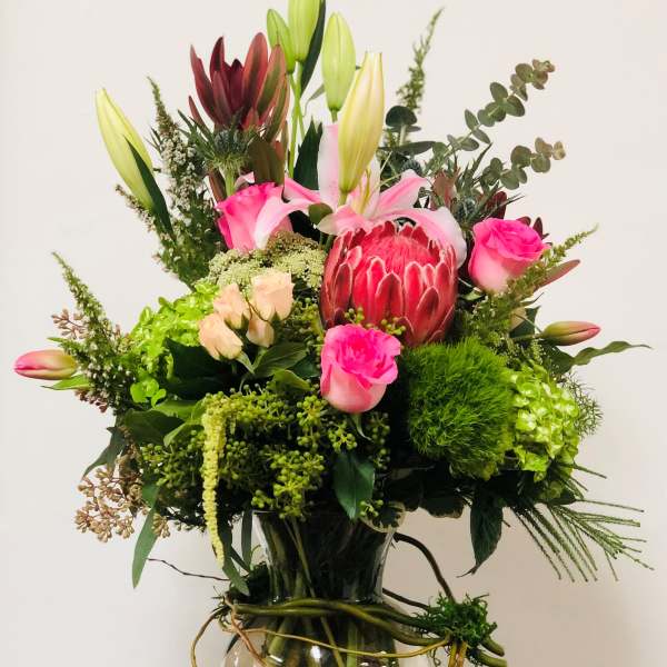 Tall mixed bouquet in a glass vase with pink lilies, roses, and a protea