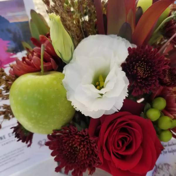 Close-up of red rose, burgundy mums, white lisianthus, and a green apple in a mixed arrangement