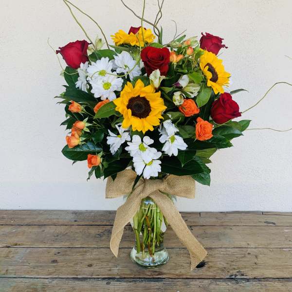 Tall mixed bouquet with sunflowers, red roses, white daisies, and orange blooms in a glass vase with burlap bow