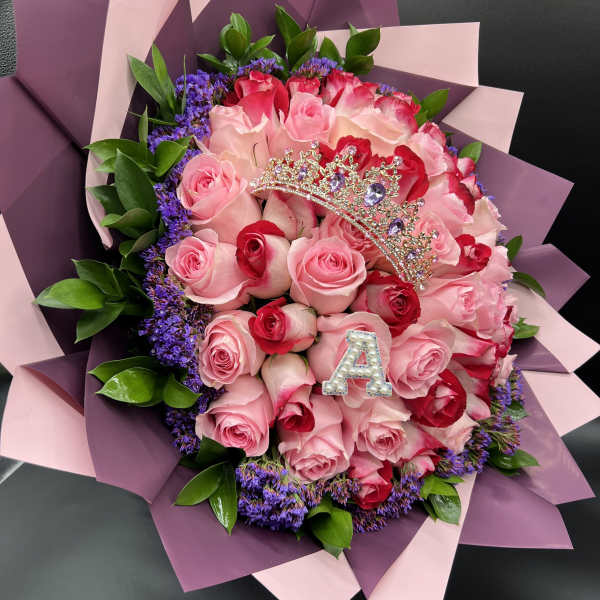 Bouquet of pink and red roses with purple filler flowers, wrapped in pink paper with a jeweled tiara and letter A.