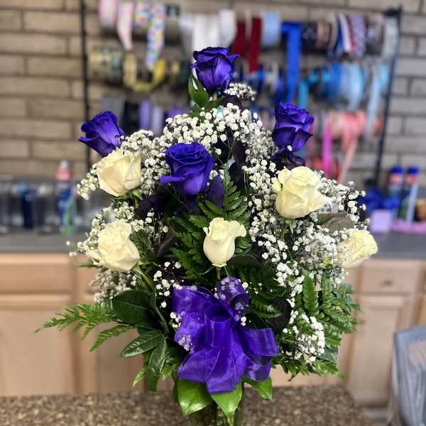 Tall arrangement of purple and white roses with baby's breath in a clear glass vase with a purple bow.