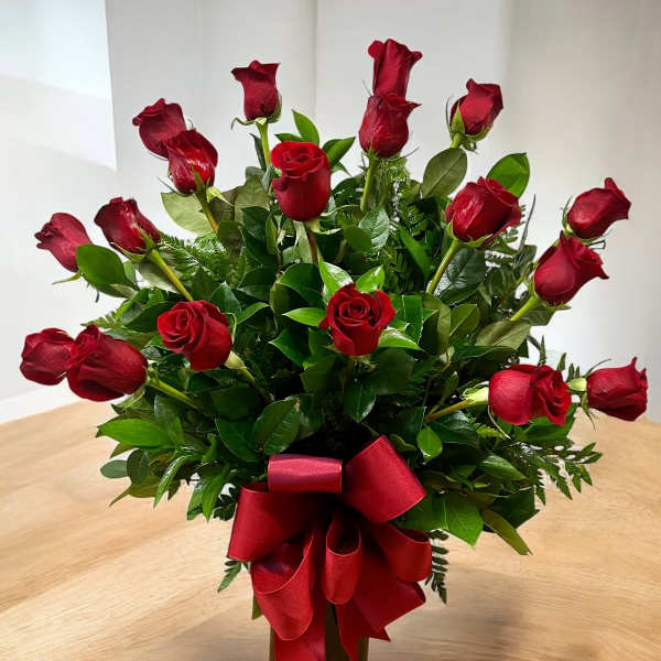 Tall arrangement of long-stem red roses with lush foliage in a dark vase with a red bow