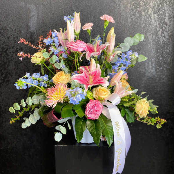 Pastel arrangement of pink lilies, roses, carnations, and blue flowers in a white vase with a white ribbon.