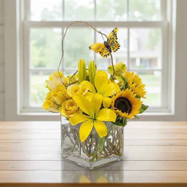 Low yellow arrangement of lilies, roses, and sunflowers in a glass vase with a decorative butterfly