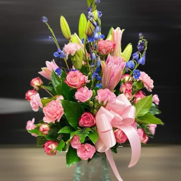 Pink roses and lilies with blue accent flowers in a glass vase with a pink bow