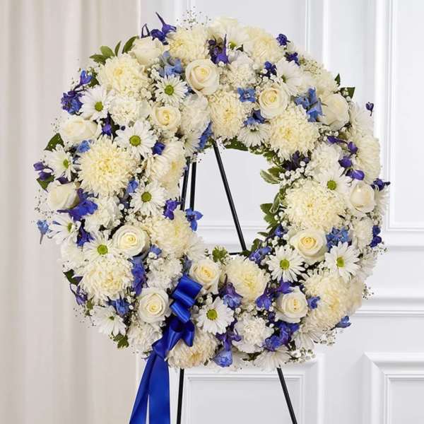 Standing wreath of white roses, daisies, and mums with blue flowers and ribbon on an easel