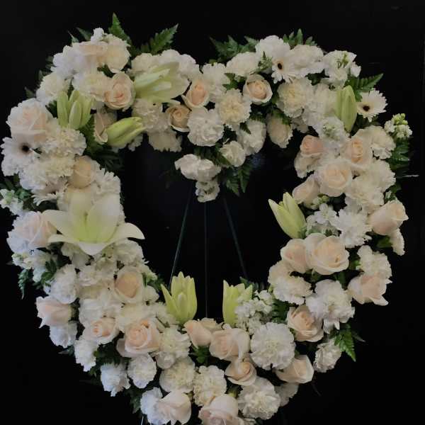 Heart-shaped white floral wreath with pale roses and lilies on a black stand