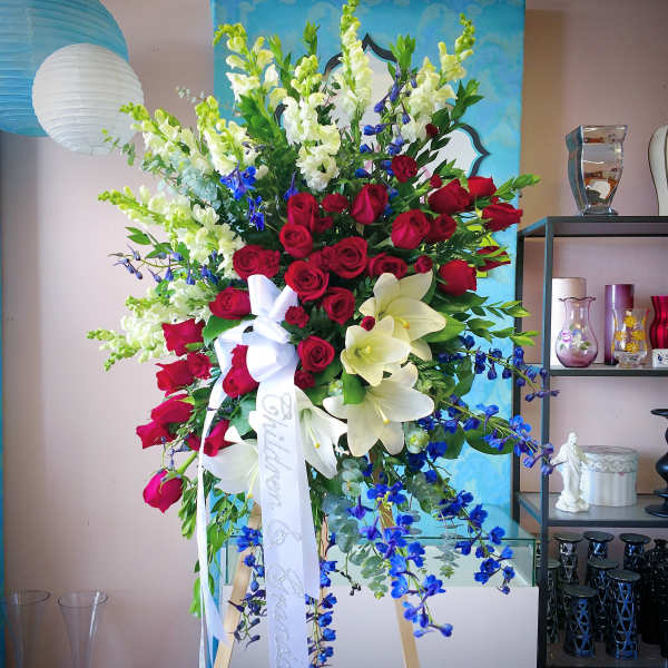 Tall floral arrangement with red roses, white lilies, and blue flowers on an easel